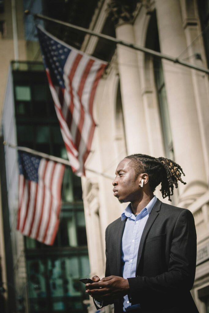 Pensive ethnic male manager standing next to building with American flags and browsing smartphone